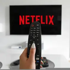 Dłoń trzymająca pilot do telewizora przed ekranem z logo Netflix.