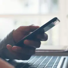 Dłoń trzymająca smartfon nad klawiaturą laptopa.