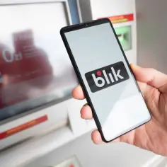 Dłoń trzyma smartfon z logo Blik. Obok bankomat, który ułatwia szybkie płatności. Kto wymyślił Blik? To polski system płatności mobilnych.