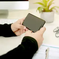 Osoba trzyma smartfon, szukając sposobu, jak zwiększyć zasięg bluetooth w telefonie. Na biurku leżą okulary i formularz.