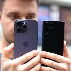 Mężczyzna trzyma dwa smartfony: fioletowy iPhone z trzema obiektywami i czarny Samsung z czterema. Oba to smartfony z dobrym aparatem, idealne do robienia zdjęć.