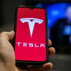 Dłoń trzyma smartfon z logo Tesli. W tle widać niewyraźną postać na niebieskim tle, być może Elon Musk. Telefon Tesla Pi.