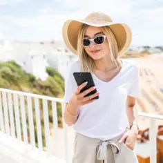 Kobieta w kapeluszu i okularach przeciwsłonecznych sprawdza telefon, ciesząc się wakacjami nad morzem. To przykład, co to roaming – możliwość korzystania z telefonu za granicą.
