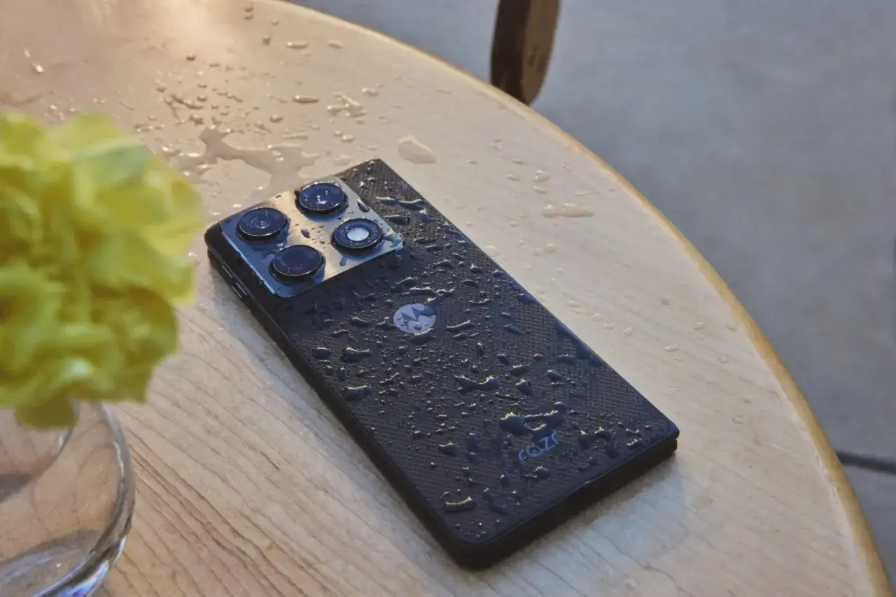 Czarny motorola razr fold z kropelkami wody na obudowie, leżący na drewnianym blacie obok żółtego kwiatu.