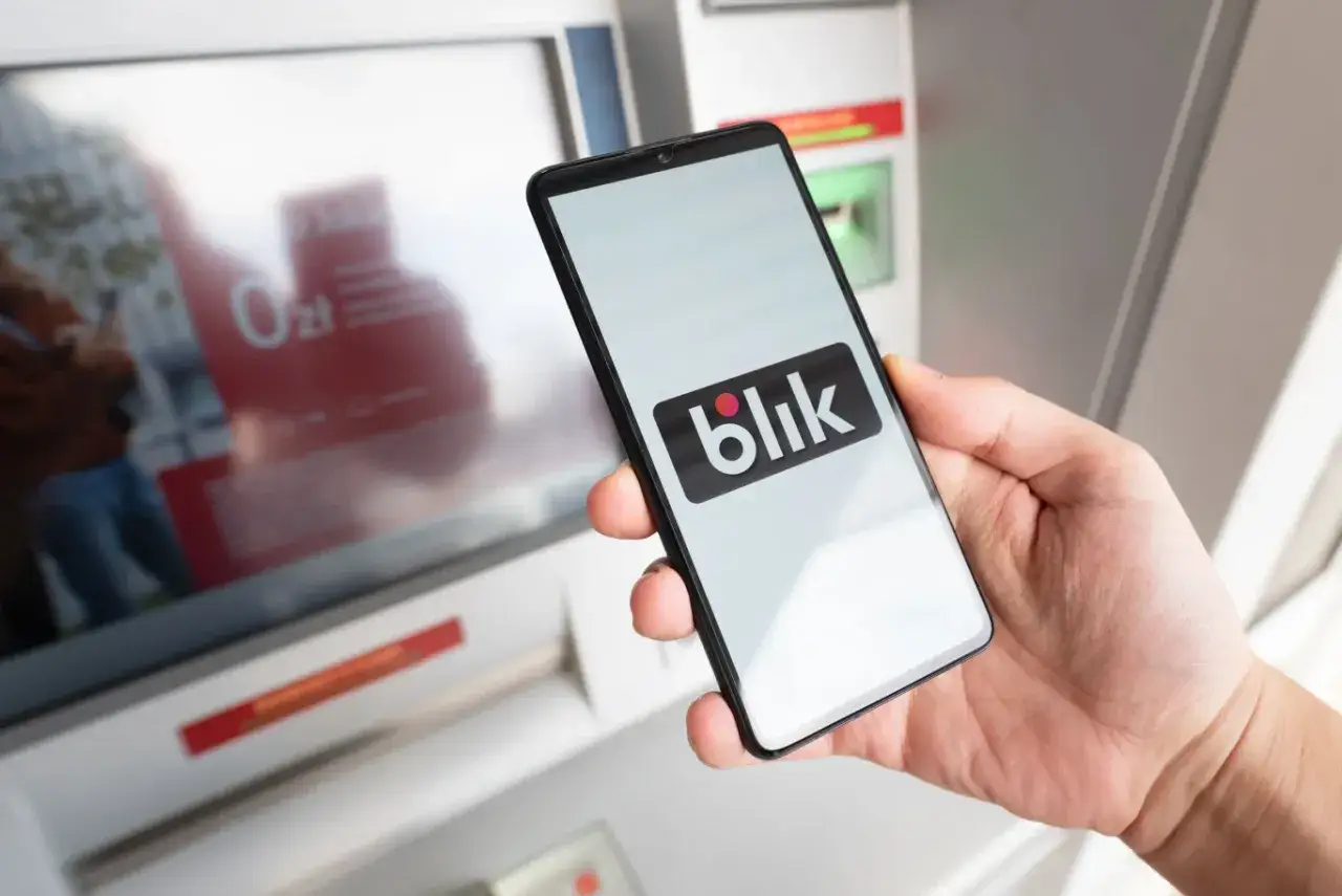 Dłoń trzyma smartfon z logo Blik przed bankomatem. Blik jest popularny w Polsce, ale jego dostępność w innych krajach jest ograniczona.