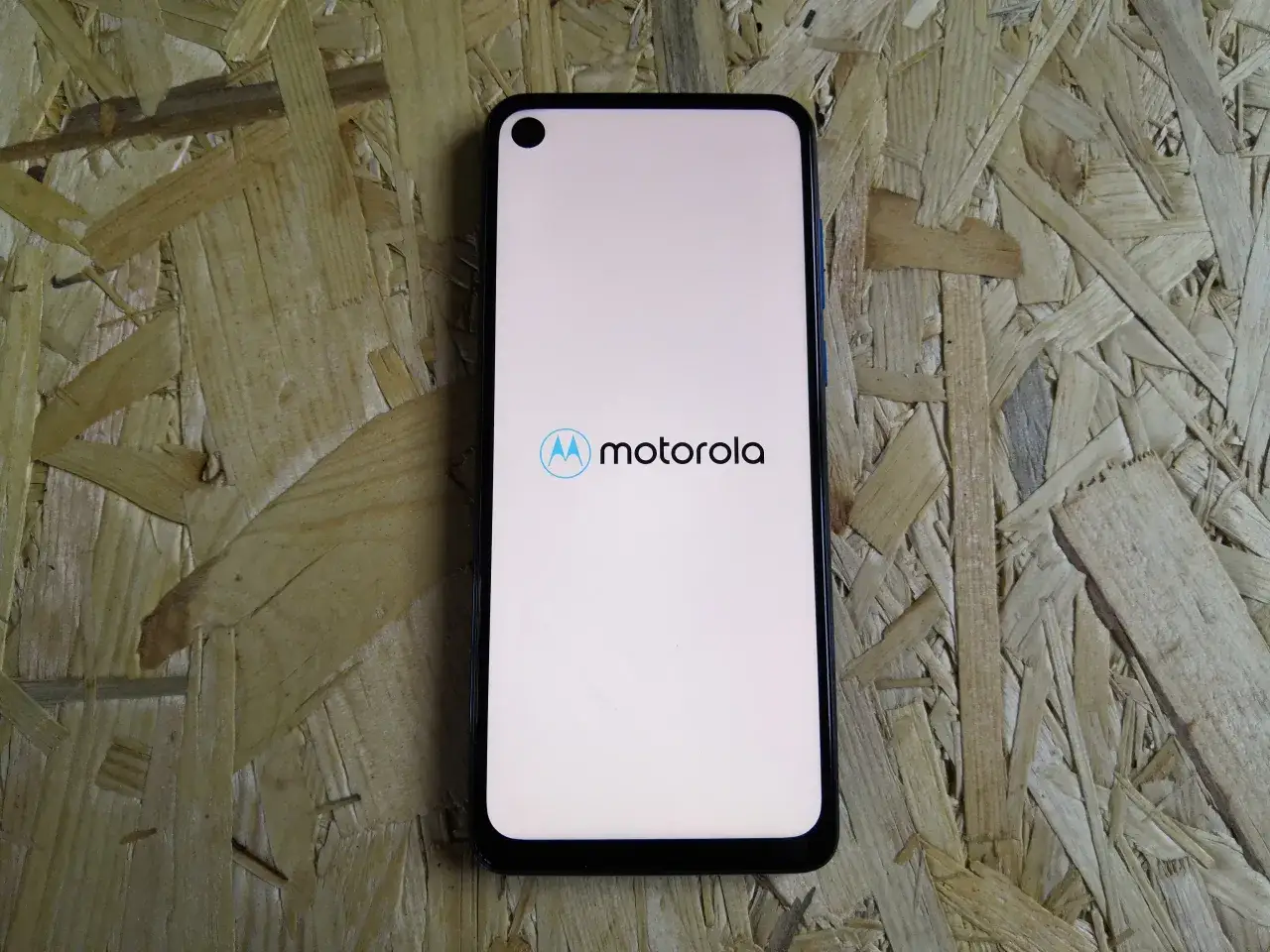 Smartfon Motorola One Vision z logo Motorola na jasnym ekranie, leżący na drewnianej płycie OSB.