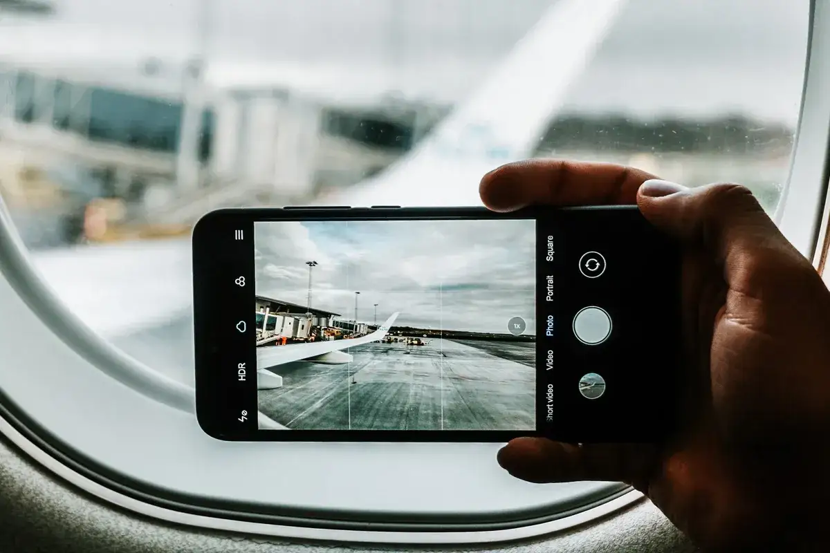 Ręka trzyma smartfon z dobrym aparatem, fotografując skrzydło samolotu i płytę lotniska przez okno samolotu.