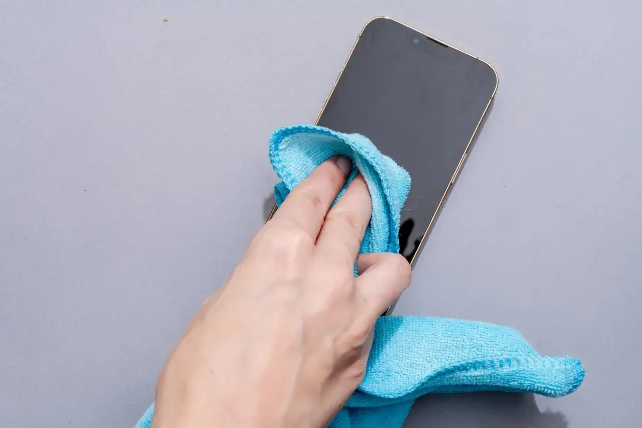 Dłoń z niebieską ściereczką z mikrofibry czyści ekran telefonu. Prosty sposób, jak wyczyścić telefon, by był jak nowy.
