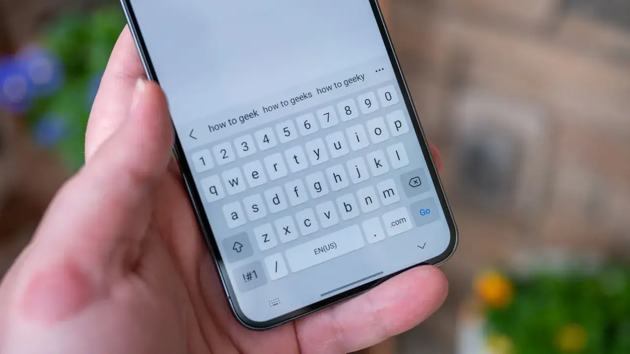 Dłoń trzyma smartfon z klawiaturą ekranową. W pasku wyszukiwania wpisano 