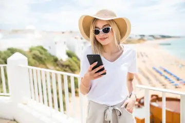 Kobieta w kapeluszu i okularach przeciwsłonecznych sprawdza telefon, ciesząc się wakacjami nad morzem. To przykład, co to roaming – możliwość korzystania z telefonu za granicą.
